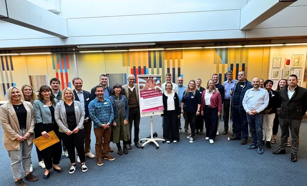 Zukunftstag Gruppe von etwa 24 Personen, formell bis lässig gekleidet, steht in einem hellen Innenraum im Halbkreis um ein auf einem Ständer platziertes Plakat.