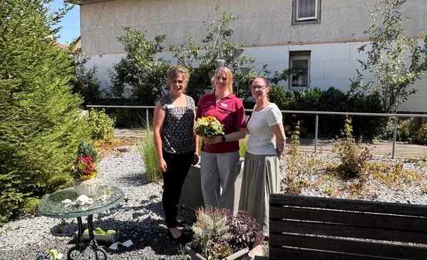 St. Wunibald Scheer Miriam Stump Drei lächelnde Frauen stehen nebeneinander in einem sonnigen Garten; die mittlere hält einen Blumenstrauß.