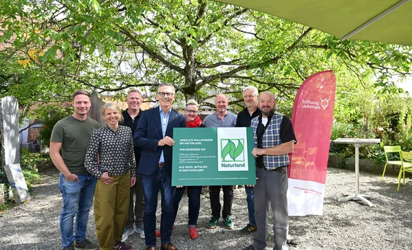 5.000ster Naturland Betrieb in Deutschland Neun lächelnde Erwachsene stehen in einem Garten unter Bäumen und halten gemeinsam ein grünes Schild mit Naturland-Logo; seitlich steht ein roter Werbebanner der Stiftung Liebenau.