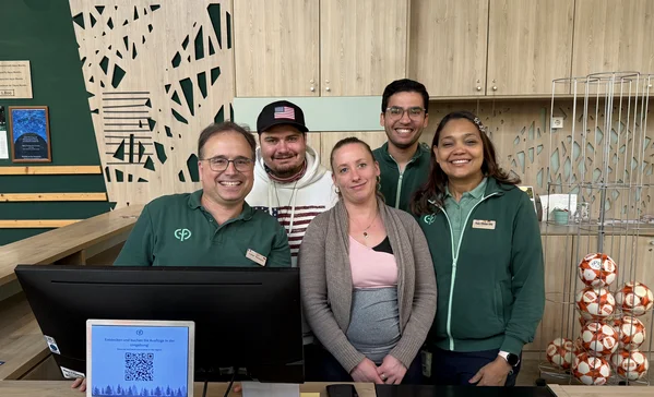 Team Rezeption Fünf lächelnde Mitarbeitende stehen eng nebeneinander hinter einer Empfangstheke; drei tragen grüne Dienstkleidung, vor ihnen ein Computermonitor.