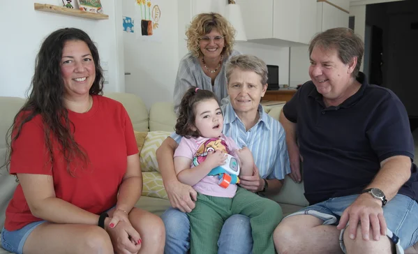 Familienunterstützender Dienst Multigenerationenfamilie auf Sofa: Eine ältere Frau hält ein kleines Mädchen auf dem Schoß, zwei Erwachsene sitzen zu beiden Seiten, eine Frau steht lächelnd dahinter.