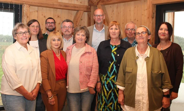 Jubilarehrung Zwölf Erwachsene posieren in einem hellen, holzgetäfelten Raum, stehend in zwei Reihen, lächelnd und gut gekleidet.