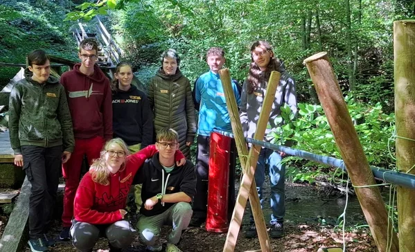 Waldtage im Berufsbildungswerk Acht Jugendliche posieren in einem Wald neben einer selbstgebauten Holzkonstruktion mit Seilen, offenbar bei einer Outdoor-Teamaktivität.
