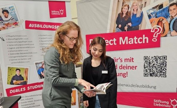 Das Bild zeigt zwei Frauen im Gespräch vor einer Wand mit Postern