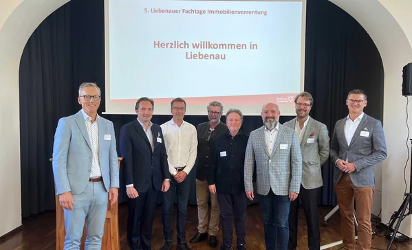 20 Jahre Immobilienverrentung in Deutschland Acht Männer in Businesskleidung stehen lächelnd vor einer Projektionsleinwand mit der Aufschrift Herzlich willkommen in Liebenau bei einer Fachveranstaltung