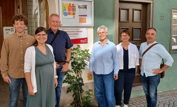 10 Jahre Ambulanten Dienste Bad Waldsee Sechs lächelnde Erwachsene unterschiedlichen Alters stehen nebeneinander vor einem Haus- bzw. Büroeingang mit Pflanzen und einem Schild mit den Worten "Inklusion" und "Ambulante Dienste