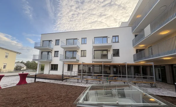 Tag der offenen Baustelle Innenhof eines modernen weißen Mehrfamilienhauses mit Balkonen, Außenterrasse mit Sitzbänken, barrierefreier Rampe und Glas-Lichtschacht im Vordergrund unter bewölktem Himmel