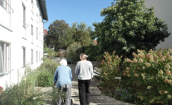 Demenzgarten Zwei Frauen gehen auf einem sonnigen Gartenweg zwischen Wohngebäuden; eine nutzt einen Rollator, die andere begleitet sie.