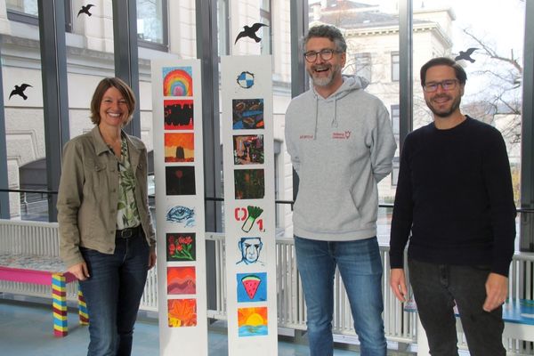 Bildungspartnerschaft Das Bild zeigt drei personen die vor der Kamera stehenund lachen. Zwischen ihnen steht eine Säule an der Plakate angebracht sind