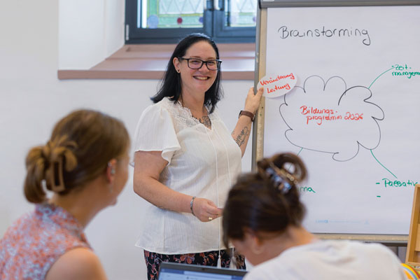 Bildungsprogramm Eine Frau zeigt an einer Tafel etwas und zwei andere Personen hören ihr zu.