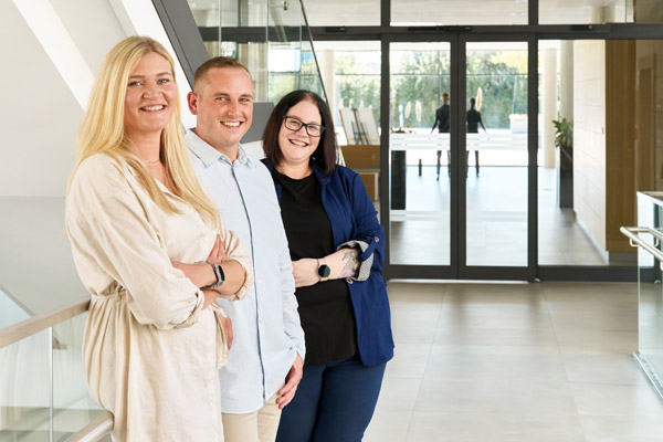 Flexteam Mitarbeitende Zwei Frauen und ein Mann stehen in einem Flur und lächeln in die Kamera.