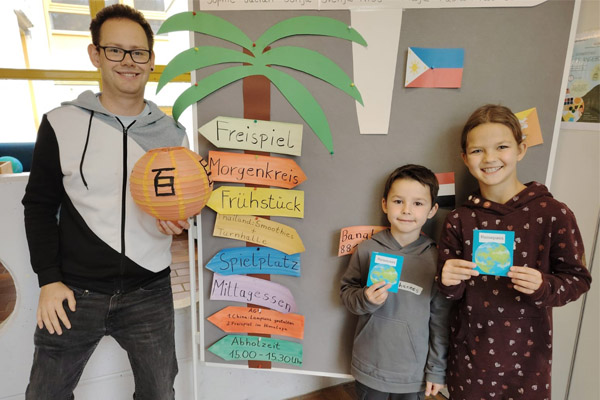 Ferienfreizeit Ein Erwachsener und zwei Kinder schauen in die Kamera und zeigen, was sie gebastelt haben.