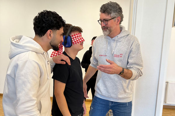 Besuch von Schülerinnen und Schülern Zwei Personen führen einen Jugendlichen mit Gehörschutz und Augenbinde durch einen Raum.