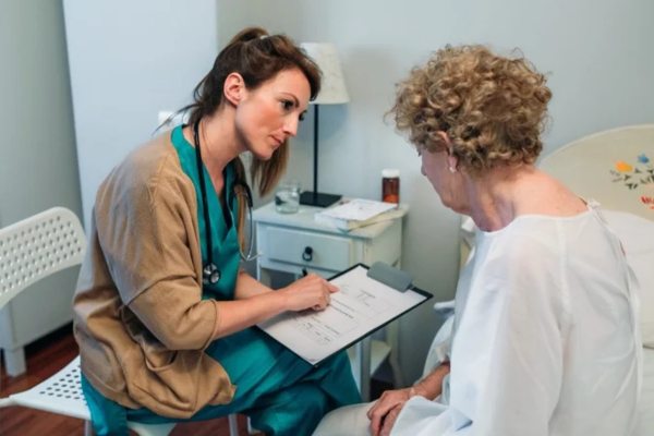 Pflegedokumentation Eine Frau mit Klemmbrett und eine Patientin in einer Unterhaltung.