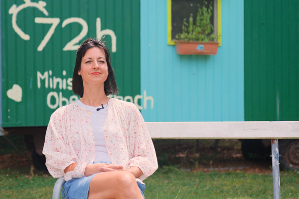 ferienbetreuung bei der stiftung liebenau das bild zeigt eine junge frau auf einer bank zum thema nachgefragt: ferienbetreuung. im hintergrund sieht man einen spiel-bauwagen.