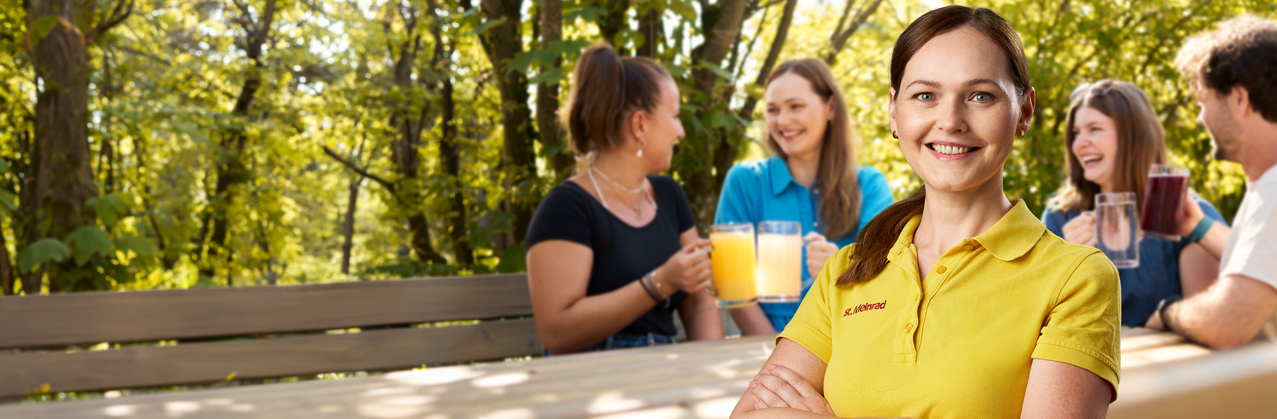 Im Vordergrund ist eine Mitarbeiterin in Arbeitskleidung zu sehen. Im Hintergrund sieht man vier junge Personen, die an einem Tisch im Garten sitzen.