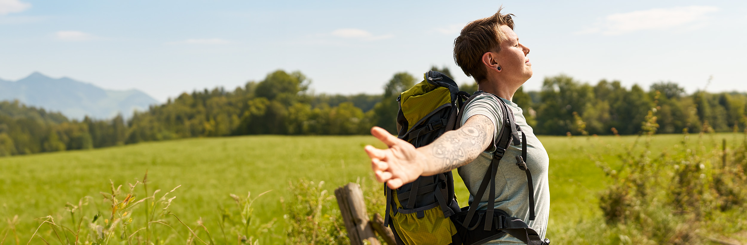 Auf dem Bild ist eine junge Frau mit ausgestreckten Armen und einem Rucksack auf einer Wiese zu sehen.