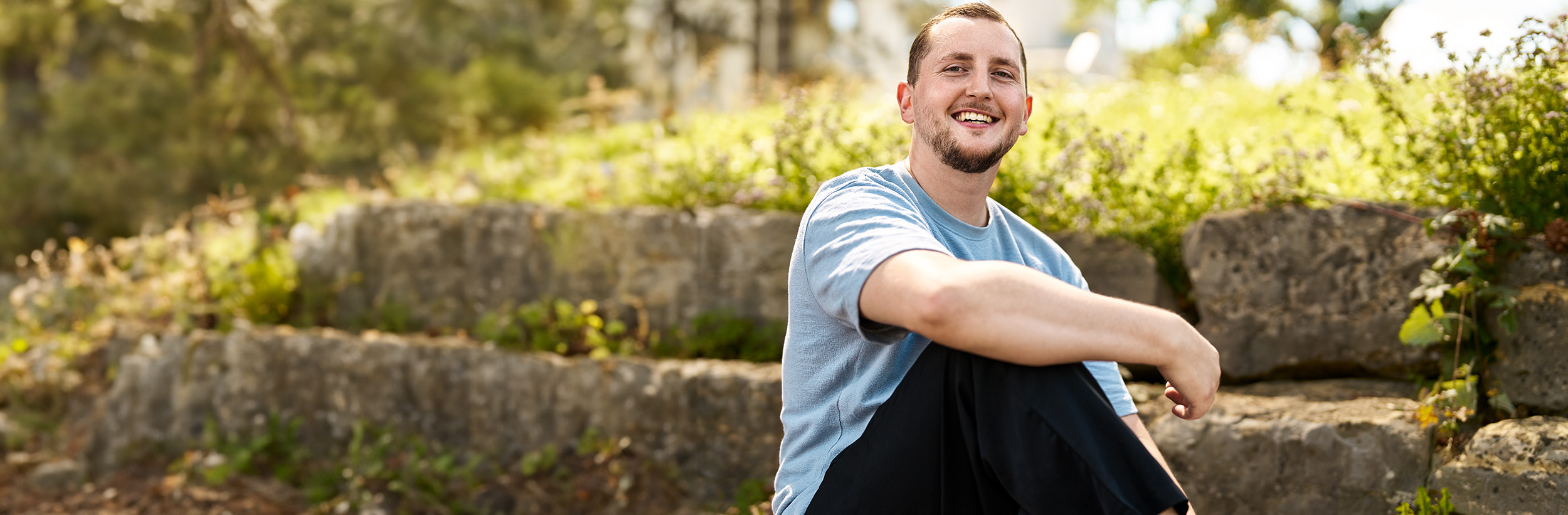 Auf dem Foto ist ein junger Mann zu sehen, der auf einer Steinmauer sitzt und lächelt.